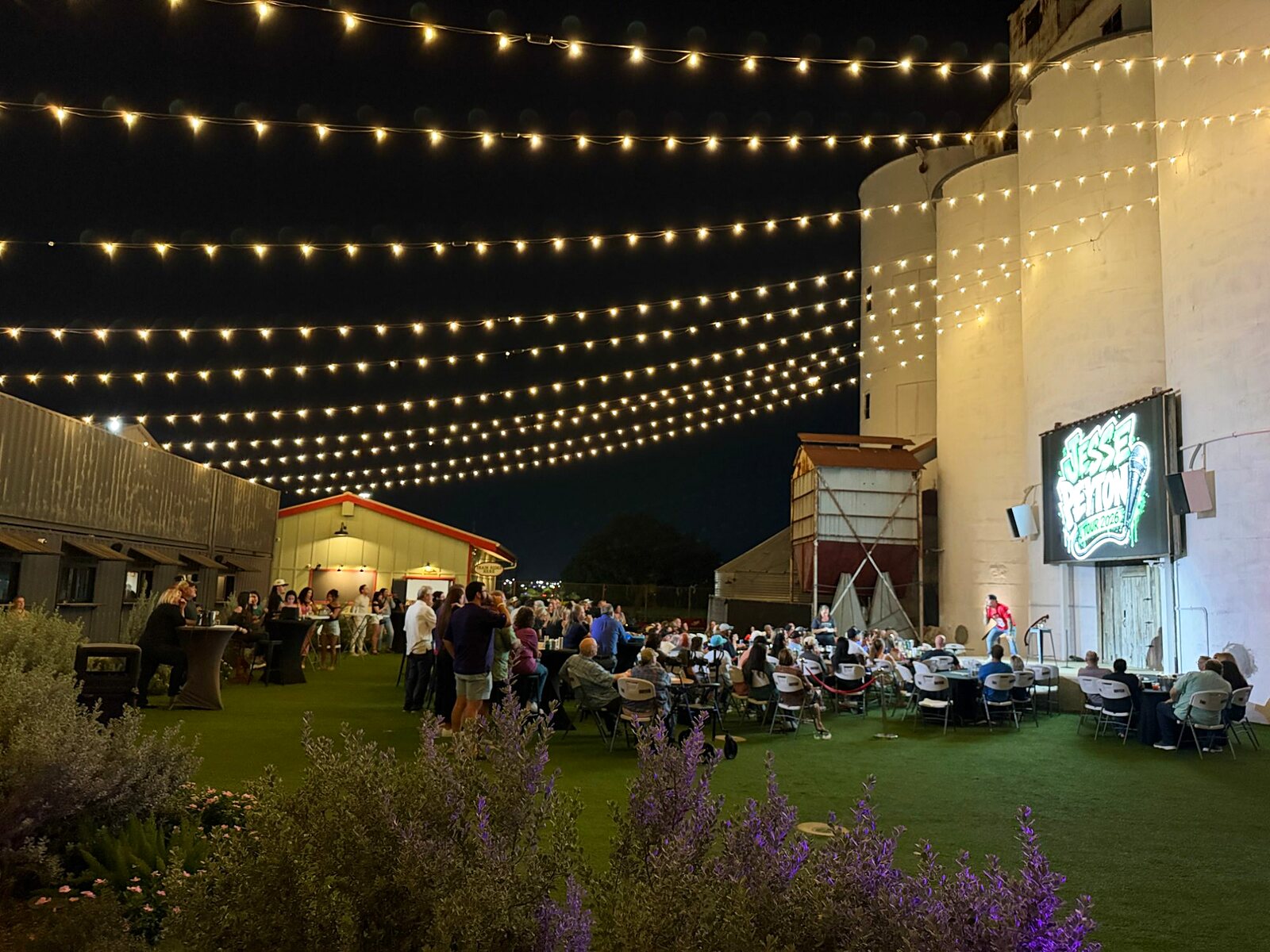 A show on the Train Yard lawn at night under string lights, the silos lit behind the stage