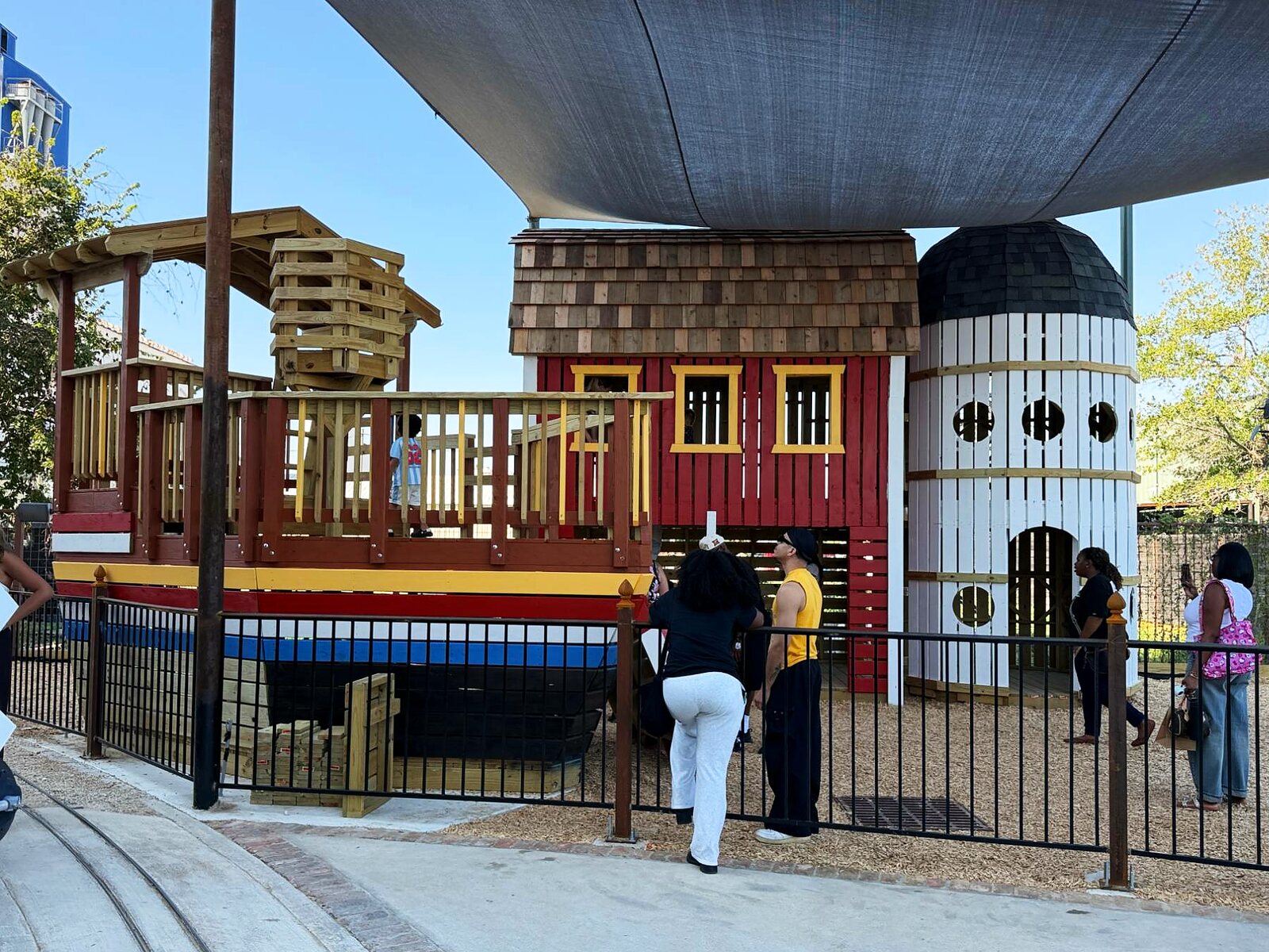 The playground: a wooden riverboat, a red barn and a white silo under a shade sail
